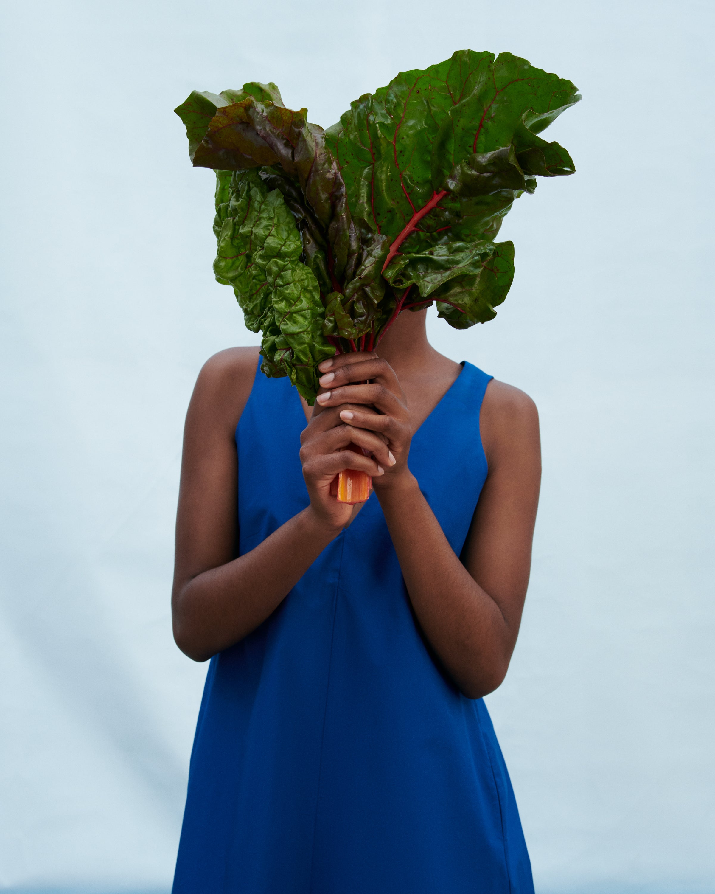A person in a bright blue sleeveless dress holds a large bunch of fresh green chard in front of their face against a pale blue studio background.