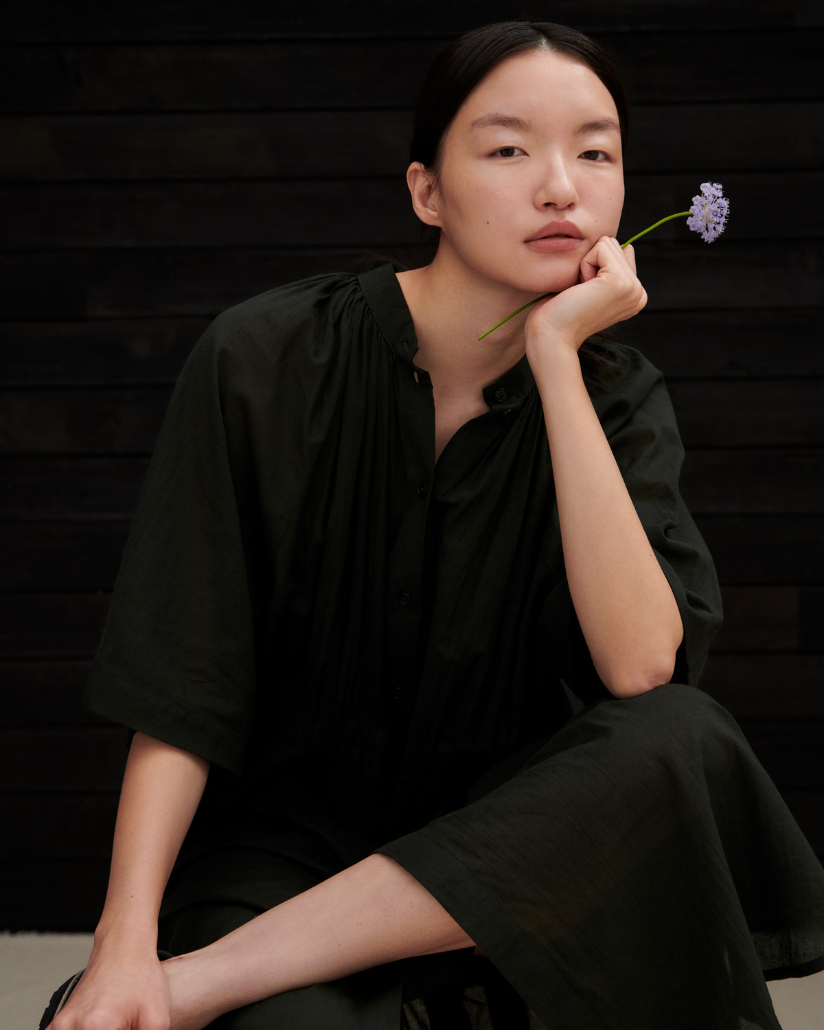 A woman wearing a loose black linen-blend top and matching pants sits against a dark wood plank background, holding a small purple flower to her chin.