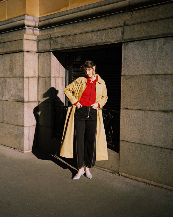 A person stands in a stone building doorway wearing a long beige trench coat over a bright red top and black trousers, with strong sunlight casting a sharp shadow on the wall.