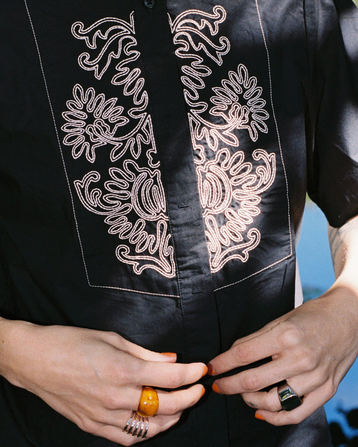 Close-up of a person wearing a black short-sleeve button-up shirt with ornate white floral embroidery on the chest, with both hands visible adjusting the front.