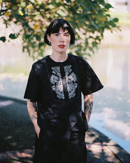 A person with short dark hair stands outdoors under leafy trees near a body of water, wearing a loose black dress with white floral embroidery around the neckline and on the lower front.