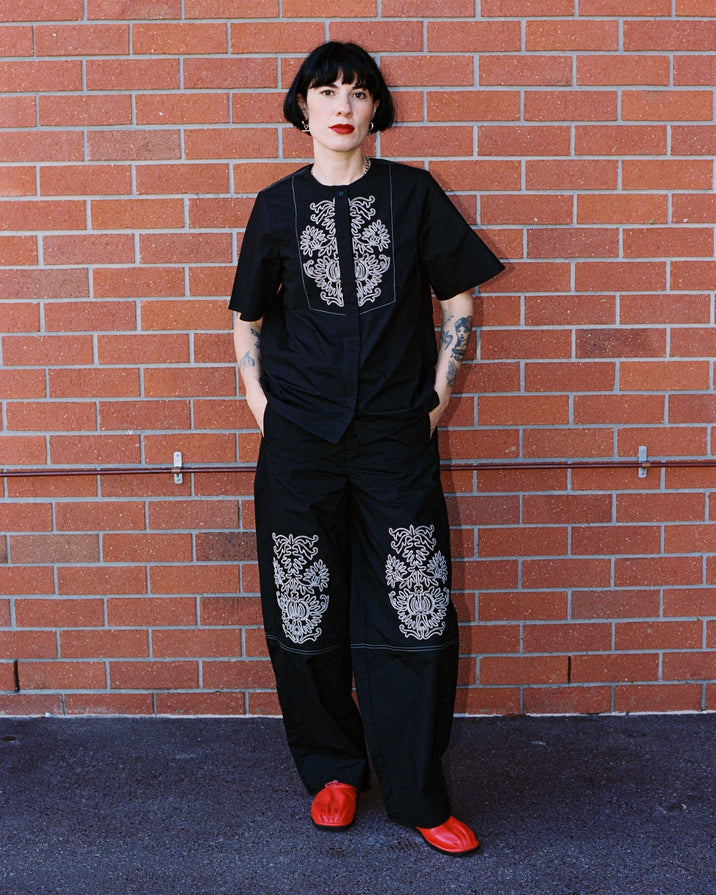 A person with short black hair and red lipstick stands against a red brick wall wearing a black short-sleeve top and matching wide-leg pants with white floral embroidery, paired with bright red shoes.
