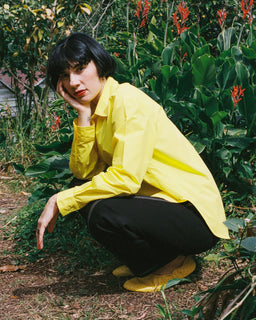 A person crouching outdoors in a garden, wearing a bright yellow button-up shirt, black pants, and yellow shoes.