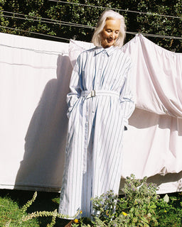 A woman with white hair stands outdoors in front of hanging white sheets, wearing a long-sleeve white shirt dress with thin blue vertical stripes and a matching belt.