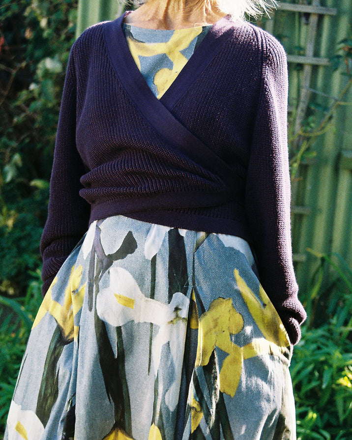 A person outdoors wearing a dark purple wrap-style knit sweater over a patterned dress featuring yellow and white floral motifs.