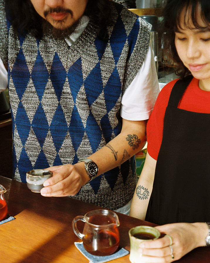 A man and a woman in a cafe setting, with the man wearing a blue, gray, and white argyle knit sweater vest over a white t-shirt. He is holding a small glass cup, while the woman in a red shirt and black apron holds a ceramic cup.