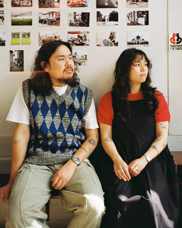Two people sitting side by side indoors, photographed against a wall covered with small photos.. The person on the left wears a white T-shirt, blue and gray argyle sweater vest, and light khaki pants.