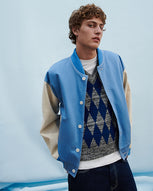 A man stands against a light blue background wearing a blue bomber jacket over a diamond patterned v neck vest and dark denim jeans.
