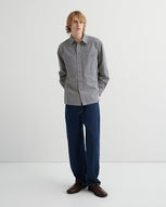 A man stands against a light grey background, wears a grey check shirt and dark denim jeans with brown shoes.