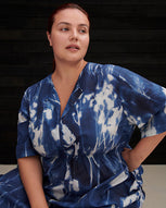 A women wearing a blue floral cyanotype print dress sitting against a black weatherboard background.