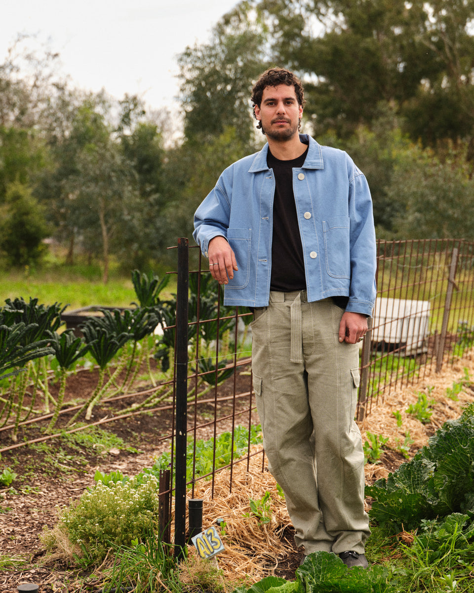 A man stands in a garden wearing a light blue denim work jacket over a black t-shirt and olive green cargo pants.