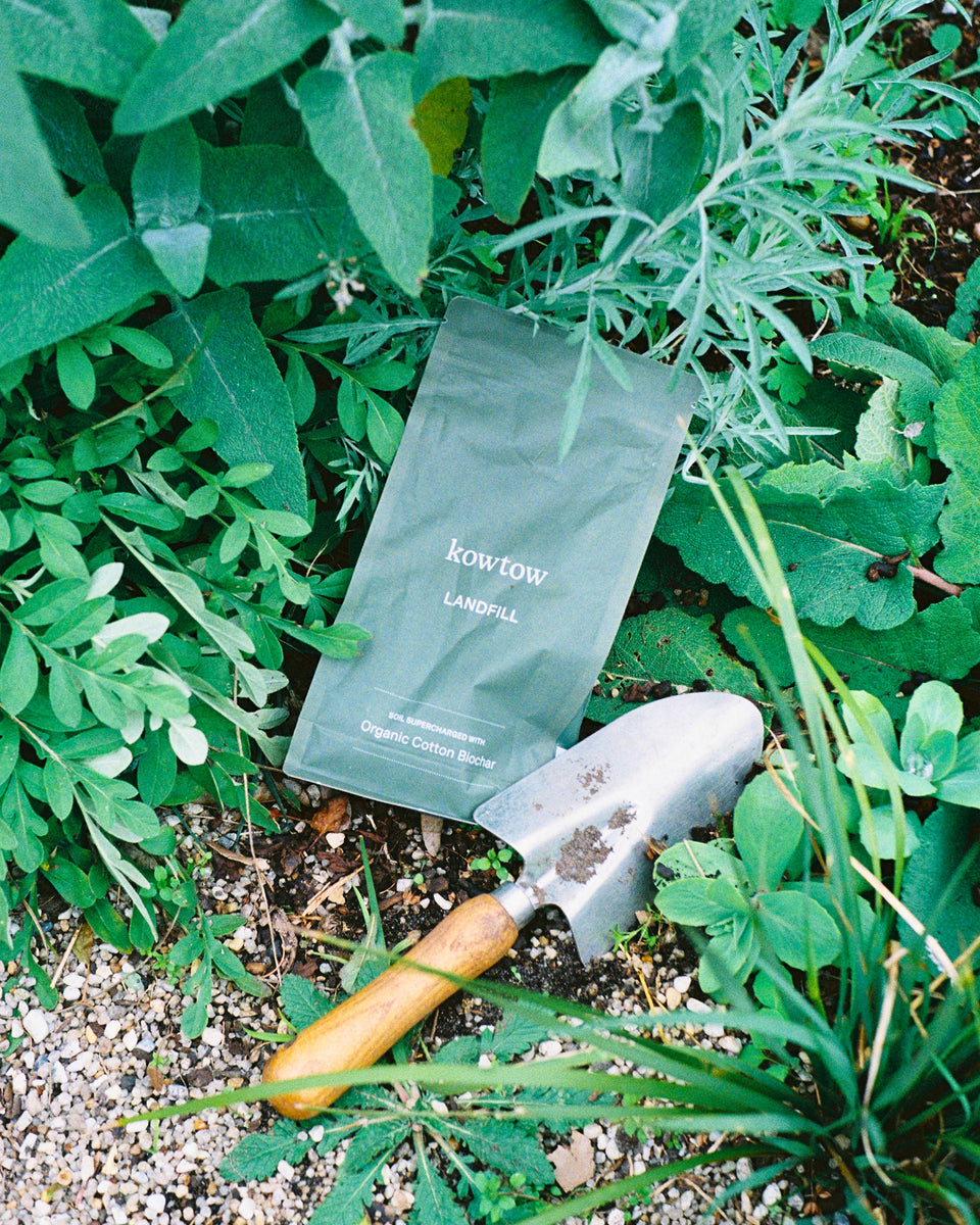 A matte dark green coffee bean bag labeled 'KOWTOW LANDFILL' lies on the ground among green plants, with a small hand trowel placed beside it.
