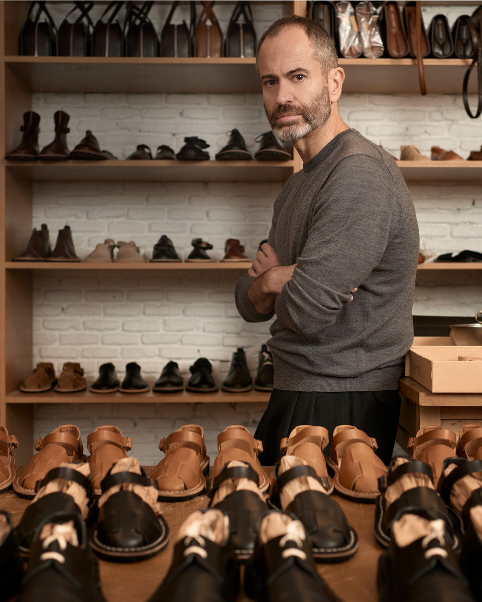 A man with a beard wearing a gray long-sleeve shirt and black trousers stands in a shoe store with shelves of shoes behind him and in the foreground.