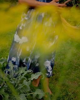 A person standing barefoot in a grassy outdoor garden, wearing a flowing blue and white tie-dye dress and holding a woven basket, with bright yellow flowers in the foreground.