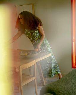 A woman with curly hair wearing a green floral print midi dress leans over a light wood dining table, placing fruit into a bowl. The photo is shot from an off-center perspective with a blurry green foreground element and warm, hazy lighting.