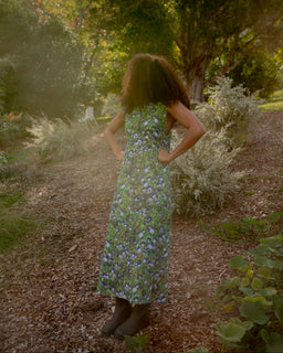 A woman stands outdoors on a wooded path wearing a sleeveless green maxi dress with a blue and purple floral print, photographed from behind.