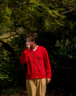 A person standing outdoors in a wooded area, wearing a bright red button up cardigan and beige pants, holding a camera.