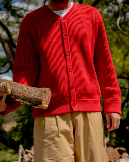 A person outdoors wearing a bright red ribbed button-front cardigan over a white undershirt and beige pants, holding a log. The background shows trees and stacked firewood in natural daylight.