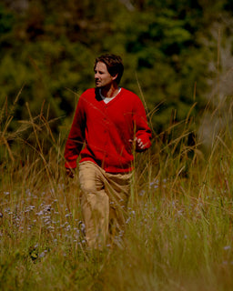 A man wearing a red knit cardigan and tan pants walking through a grassy outdoor field with trees in the background.