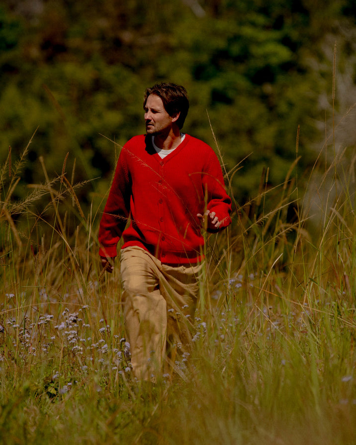 A man wearing a red knit cardigan and tan pants walking through a grassy outdoor field with trees in the background.