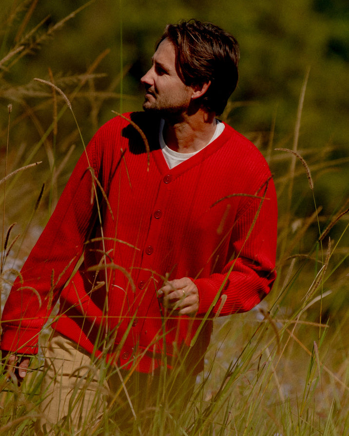 A man wearing a bright red knit cardigan over a white undershirt and tan pants, standing in tall sunlit grass outdoors.