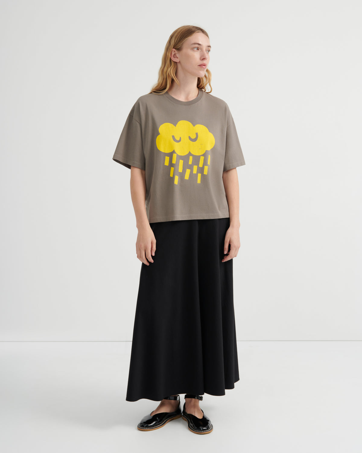 A woman modeling a taupe short-sleeve t-shirt with a yellow cloud and rain graphic, paired with a long black skirt and black shoes, against a plain white studio background.