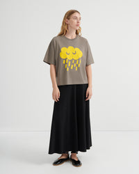 A woman modeling a taupe short-sleeve t-shirt with a yellow cloud and rain graphic, paired with a long black skirt and black shoes, against a plain white studio background.