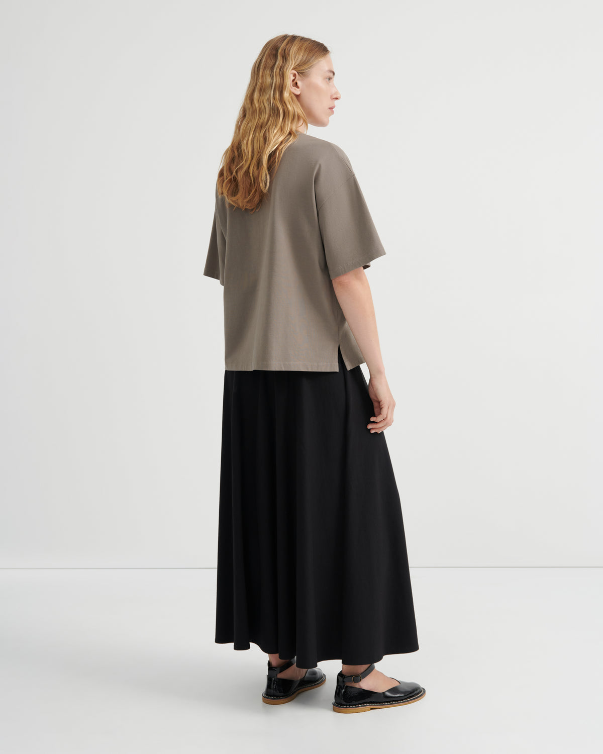 A woman stands in profile wearing an oversized taupe short-sleeve top and a long black skirt, photographed in a clean studio setting.