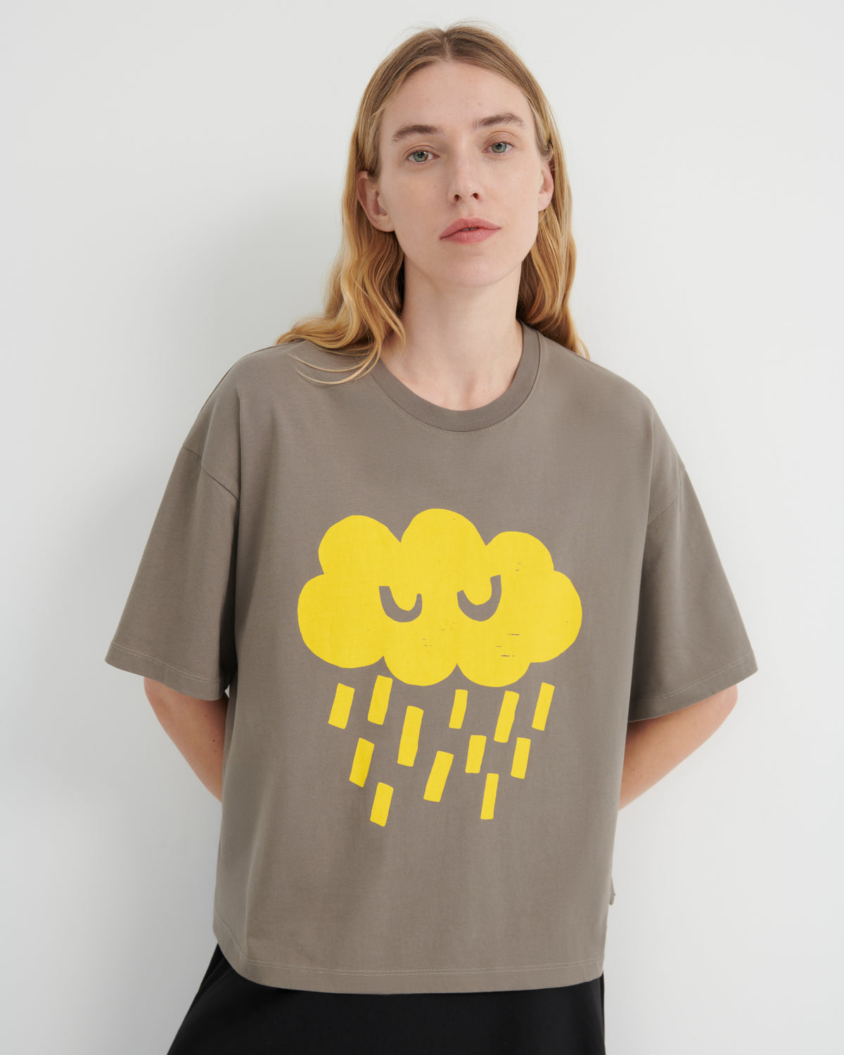 A woman wearing a taupe-gray oversized t-shirt with a yellow cartoon cloud and raindrop graphic on the front, set against a plain light gray background.