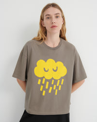 A woman wearing a taupe-gray oversized t-shirt with a yellow cartoon cloud and raindrop graphic on the front, set against a plain light gray background.