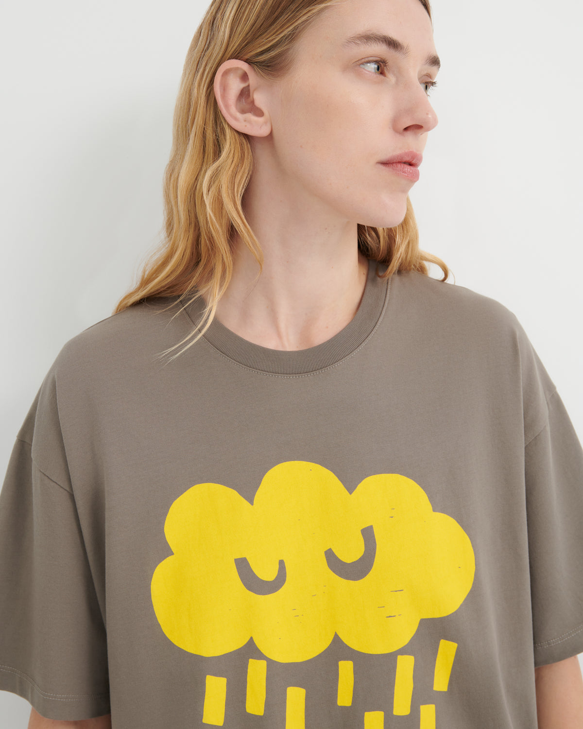 A woman wearing an oversized taupe-gray t-shirt with a large yellow cartoon cloud graphic on the front, shown in a close-up profile view against a light gray background.