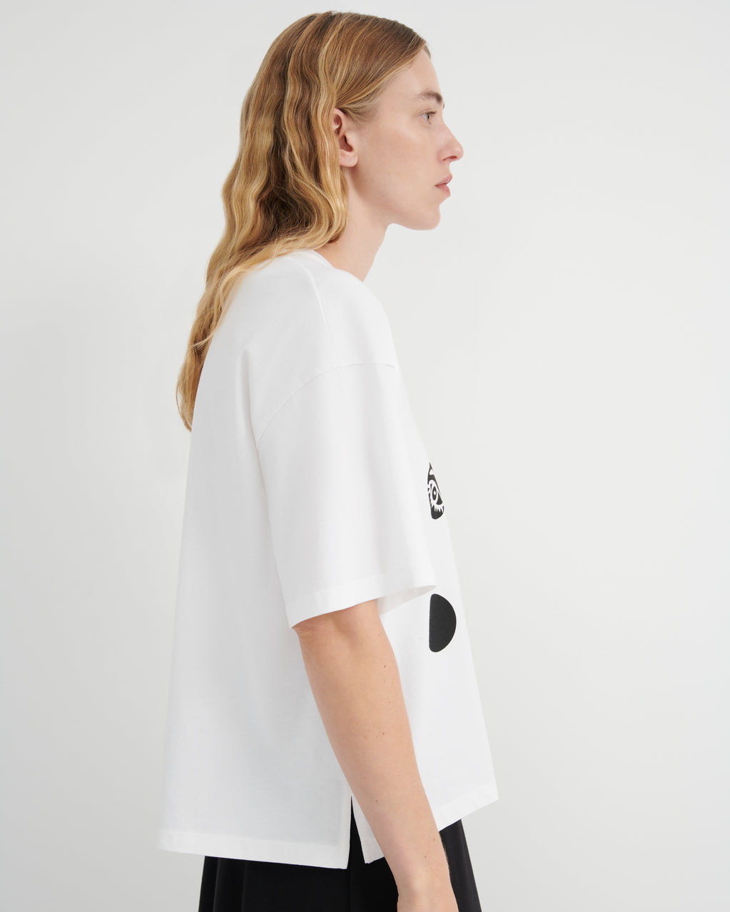 A woman shown in profile wearing an oversized white T-shirt with black graphic accents on the side, photographed against a plain light gray studio background.
