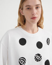A close-up of a model wearing a white crewneck sweatshirt with black graphic prints, including solid circles and stylized eyes, against a plain light background.