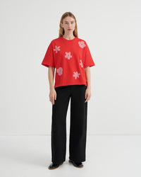 A woman stands against a plain light gray studio background wearing a bright red short-sleeve T-shirt with large pink flower motifs and black wide-leg trousers.