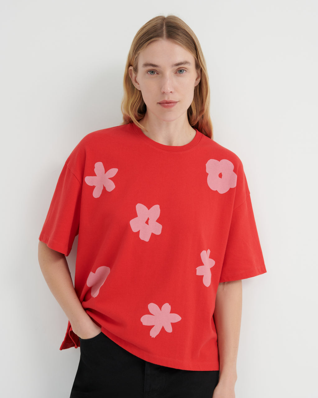 A woman wearing a bright red oversized short-sleeve t-shirt with light pink flower motifs, paired with black pants, set against a plain light gray background.