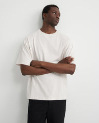 A man wearing an oversized off-white short-sleeve t-shirt and black pants, standing with arms crossed.