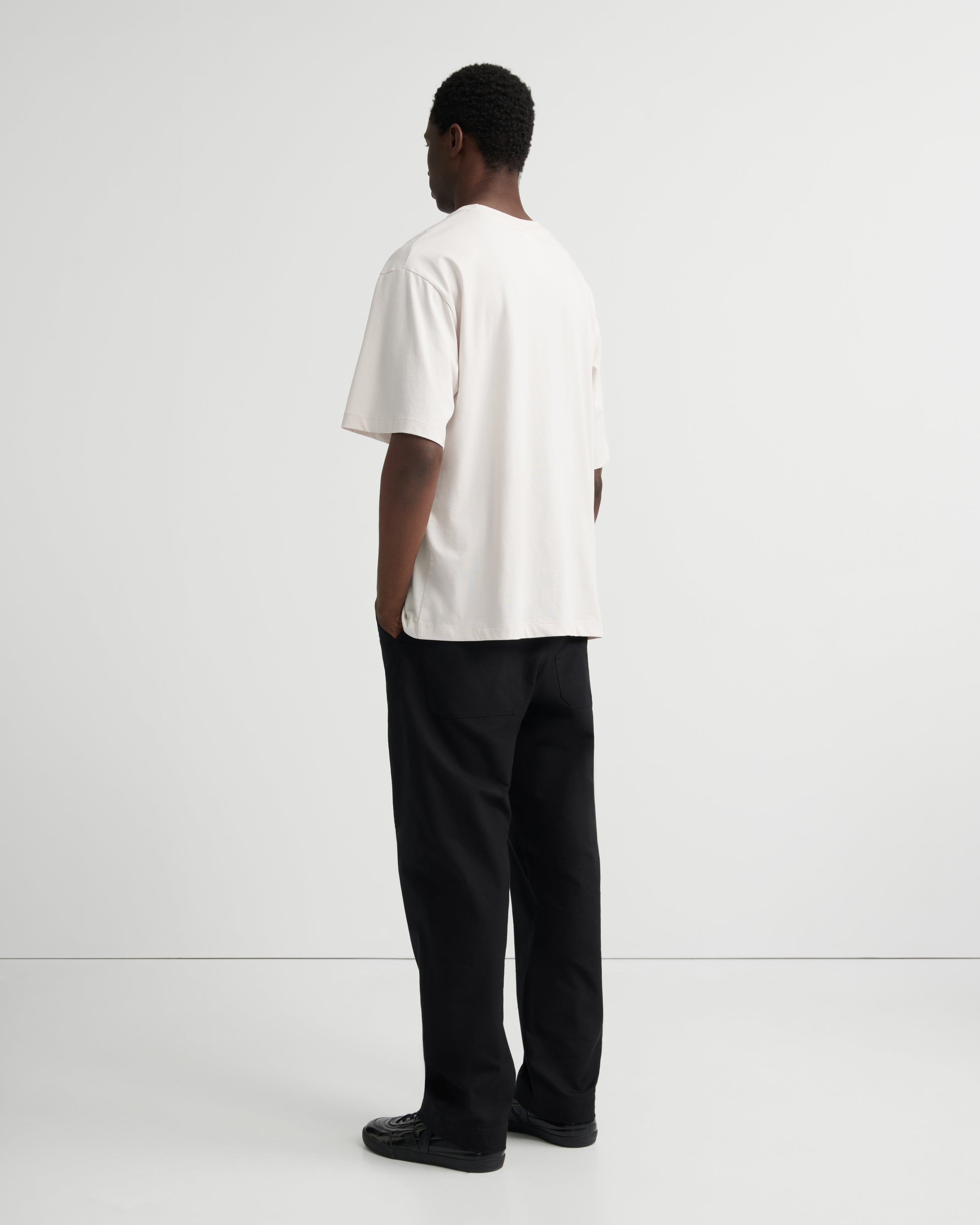 A man standing with his back to the camera, wearing a loose white short-sleeve t-shirt and black trousers.