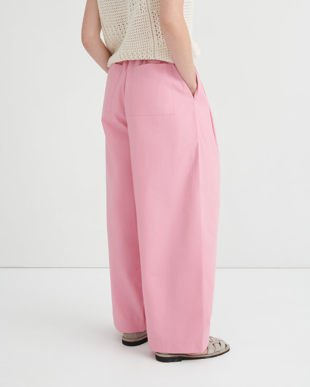 A model shown from the back wearing pink wide-leg trousers, a cream knit top, and flat sandals against a plain white studio background.