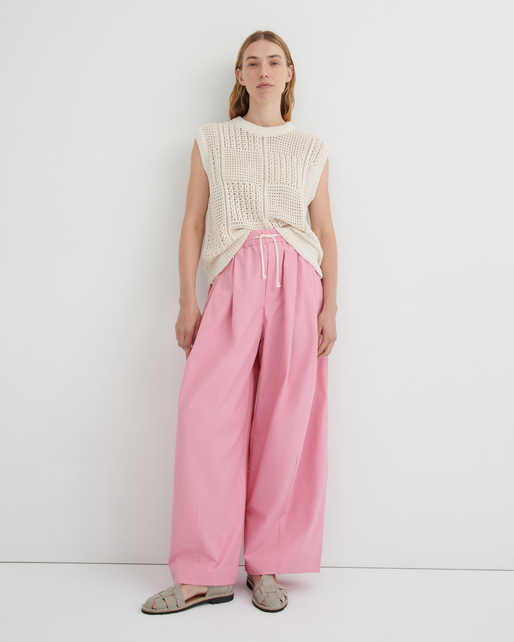 A model stands against a plain white studio background wearing a cream sleeveless knit top, pink wide-leg drawstring pants, and beige flat sandals.