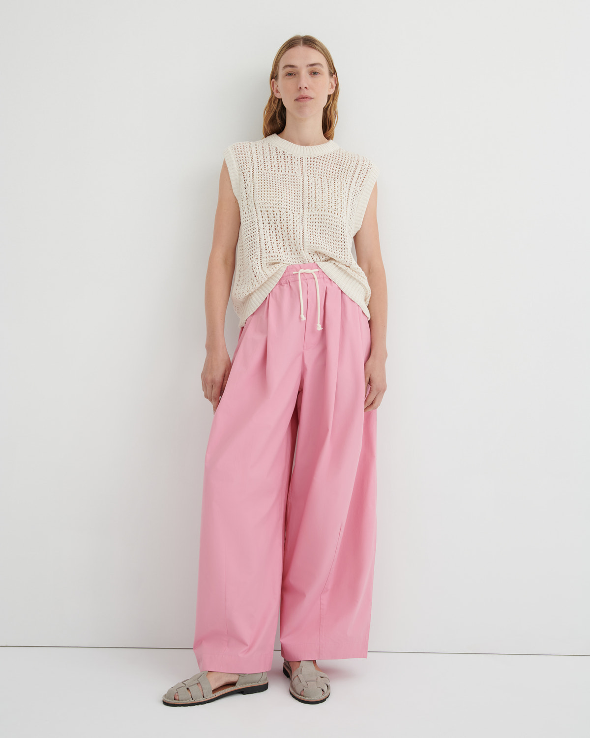 A model stands against a plain white studio background wearing a cream sleeveless knit top, pink wide-leg drawstring pants, and beige flat sandals.