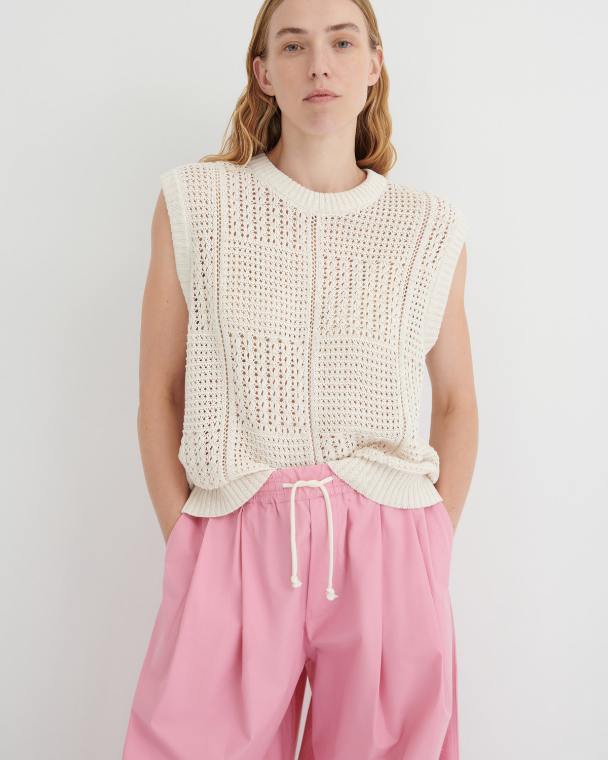 A model wearing a cream-colored sleeveless open-knit sweater vest and pink drawstring trousers, standing against a light gray studio background.