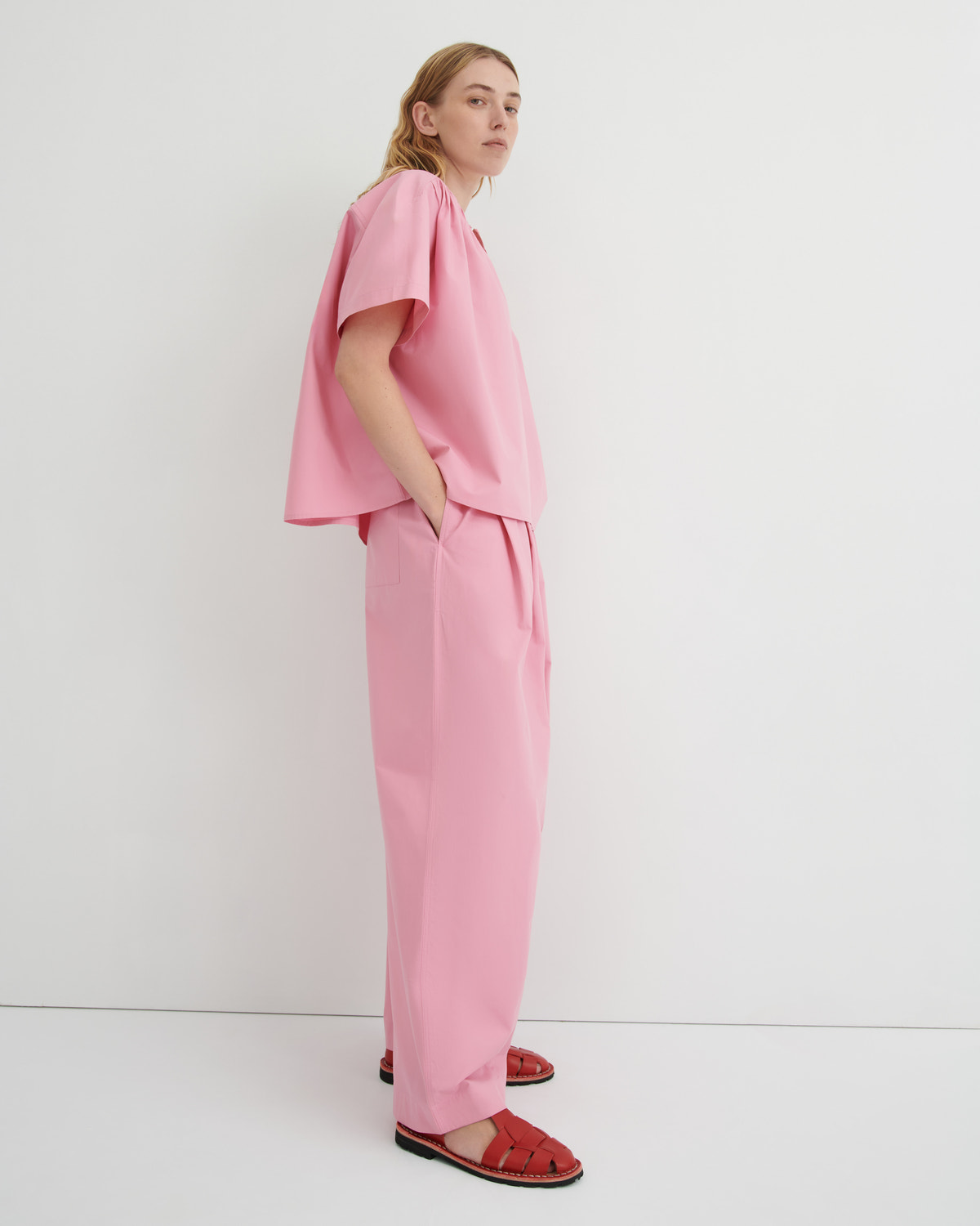A model stands in profile wearing a matching pink two-piece outfit consisting of a short-sleeve top with voluminous draped sleeves and wide-leg trousers, paired with red sandals against a clean white studio background.