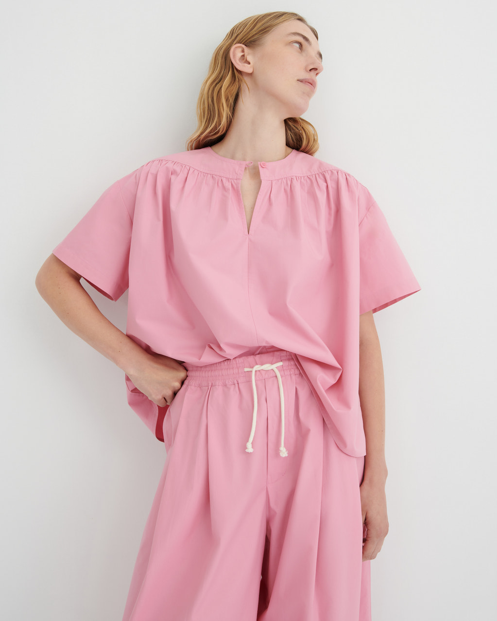 A woman wearing a pink short-sleeve top with a gathered neckline and keyhole cutout, paired with matching pink drawstring pants, against a plain light gray studio background.