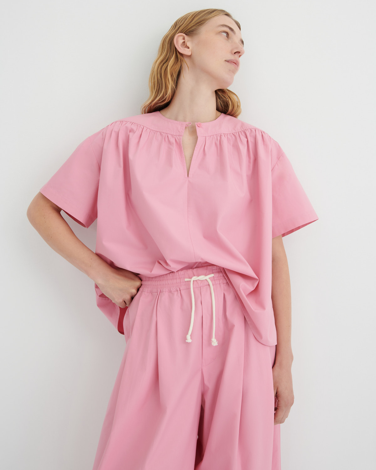 A woman wearing a pink short-sleeve top with a gathered neckline and keyhole cutout, paired with matching pink drawstring pants, against a plain light gray studio background.