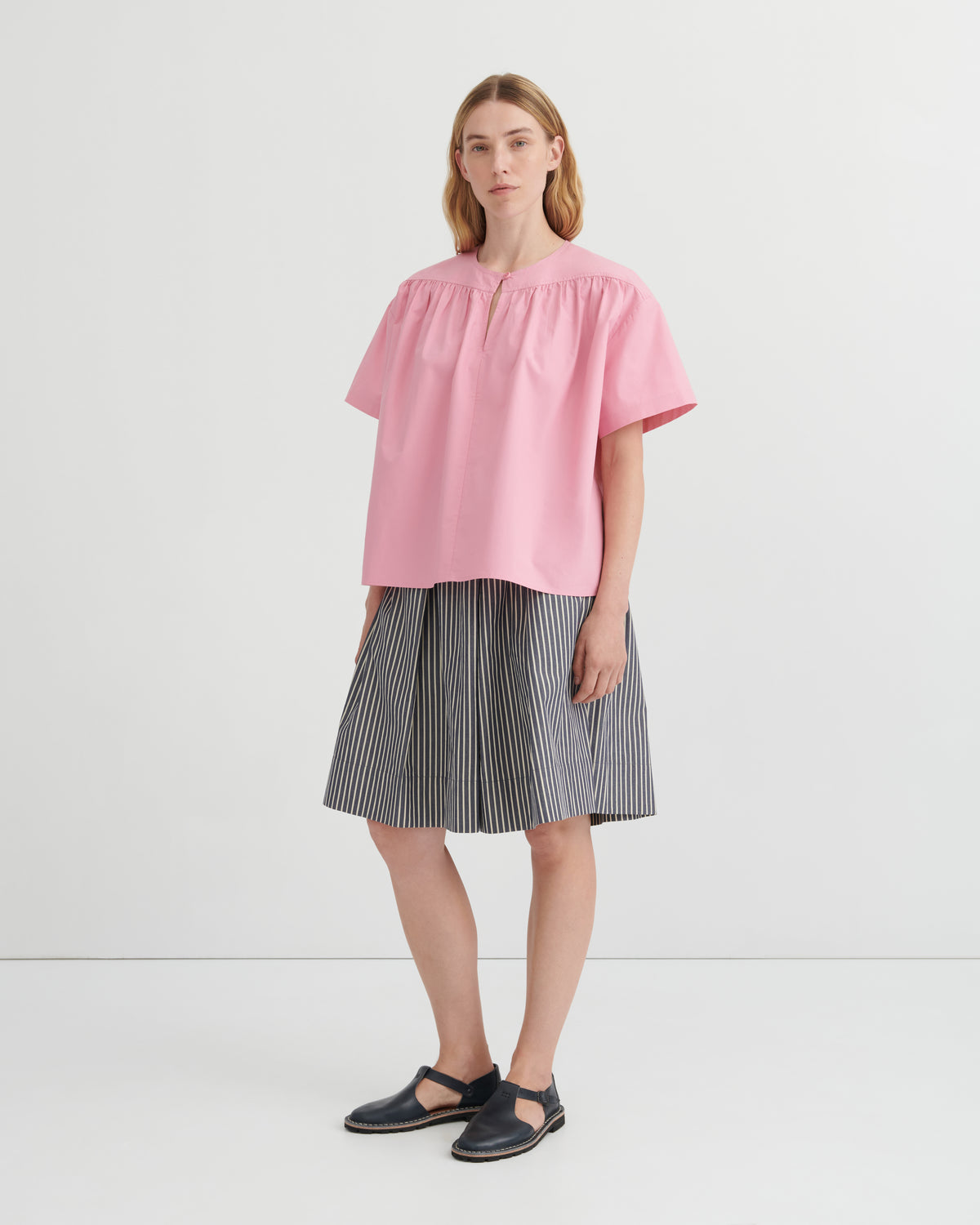 A woman modeling a light pink short-sleeve blouse with a gathered neckline and pleated skirt in a studio setting.