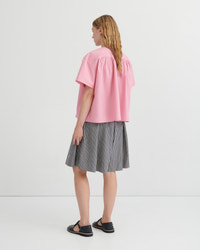 A woman shown from the back wearing a pink short-sleeve blouse with gathered shoulders and a gray-and-white vertically striped pleated skirt, paired with black flat sandals, against a plain light gray studio background.