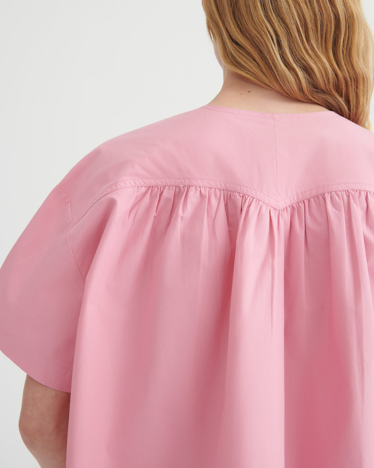 Close-up back view of a person wearing a loose pink short-sleeve top with gathered shoulder seams, against a plain light background.