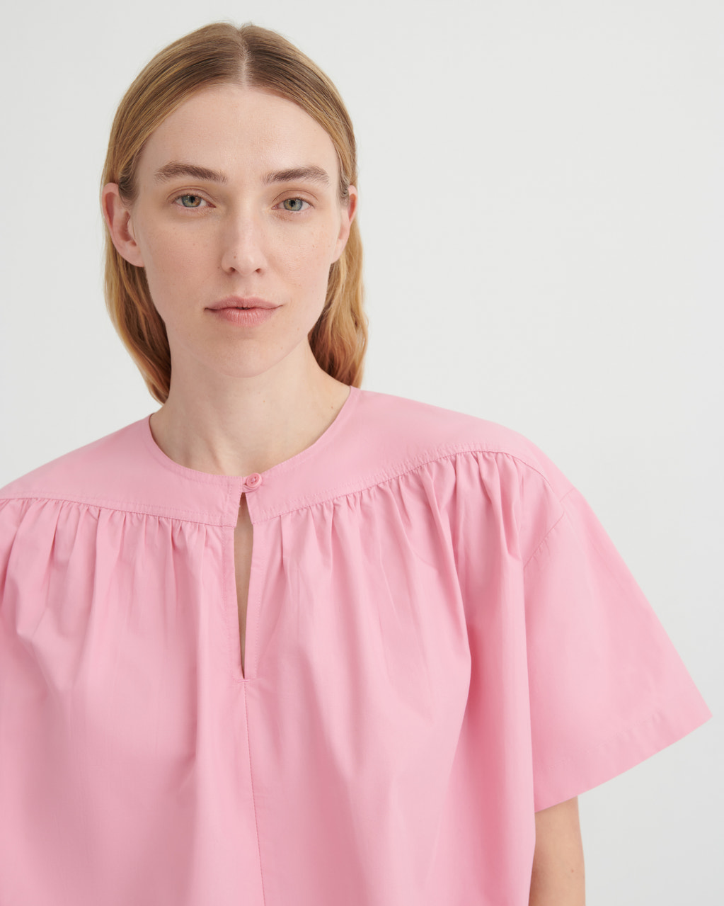 A woman wearing a light pink short-sleeve blouse with a gathered round neckline and a small front keyhole cutout, photographed against a plain light gray studio background.