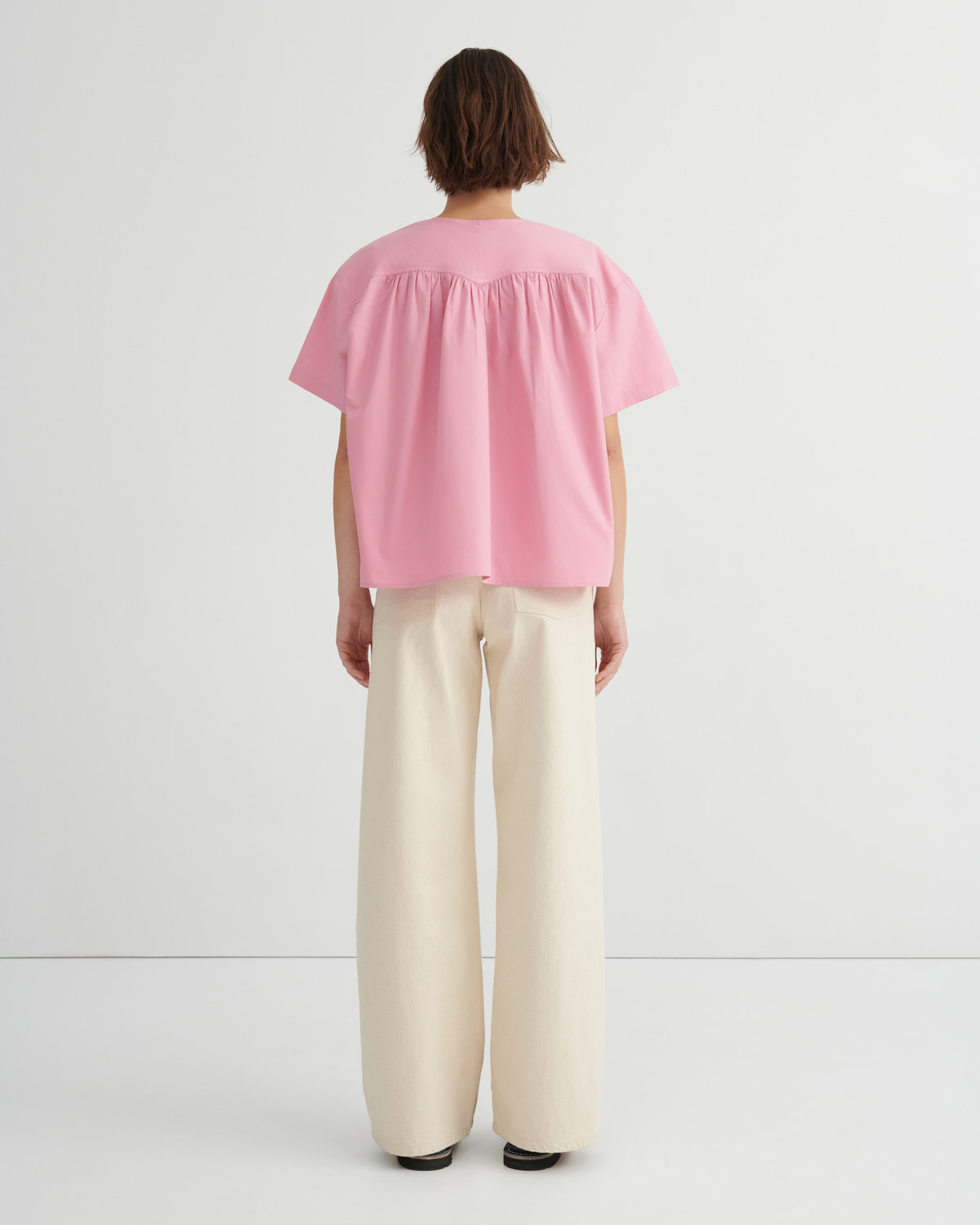 A model shown from the back wearing a light pink short-sleeve blouse with a gathered, ruffled neckline and wide-leg cream trousers, set against a plain white studio background.