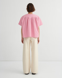 A model shown from the back wearing a light pink short-sleeve blouse with a gathered, ruffled neckline and wide-leg cream trousers, set against a plain white studio background.